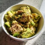 A close-up of a white bowl filled with creamy Avocado Tuna Salad, seasoned with black pepper.