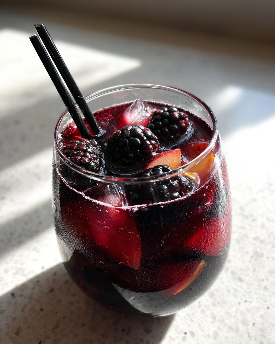 A refreshing glass of Black Magic Sangria for Halloween, filled with dark berries, fruit slices, and ice, with black straws.