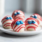 Close-up of several white chocolate-covered truffles decorated to look like mummies with red icing and blue M&M eyes, representing Bloody Eyeball Truffles.