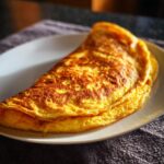 A perfectly cooked golden omelet, folded on a white plate, ready to eat. Simple and delicious omelet.