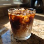 Close-up of a glass filled with iced caramel macchiato coffee, showing swirls of cream and dark coffee over ice.