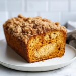 A close-up of a freshly baked Pumpkin Streusel Bread loaf on a white plate, showcasing its crumbly topping and moist interior.