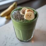 A vibrant green spinach smoothie in a glass, topped with banana slices and chia seeds.