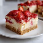 A close-up of a Strawberry Shortcake Cheesecake Bar slice on a white plate, topped with fresh strawberries and strawberry sauce.