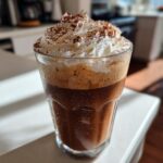 A tall glass of Tiramisu Iced Coffee topped with whipped cream and dusted with cocoa powder.