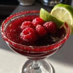 A festive Vampire's Kiss Martini cocktail garnished with fresh raspberries and lime wedges, with a red sugar rim.