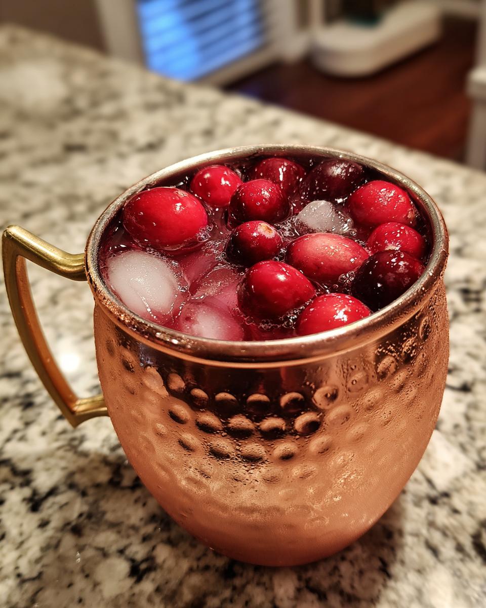 A close-up of a festive cranberry cocktail in a hammered copper mug, garnished with fresh cranberries and ice. One of the best Thanksgiving cocktails.