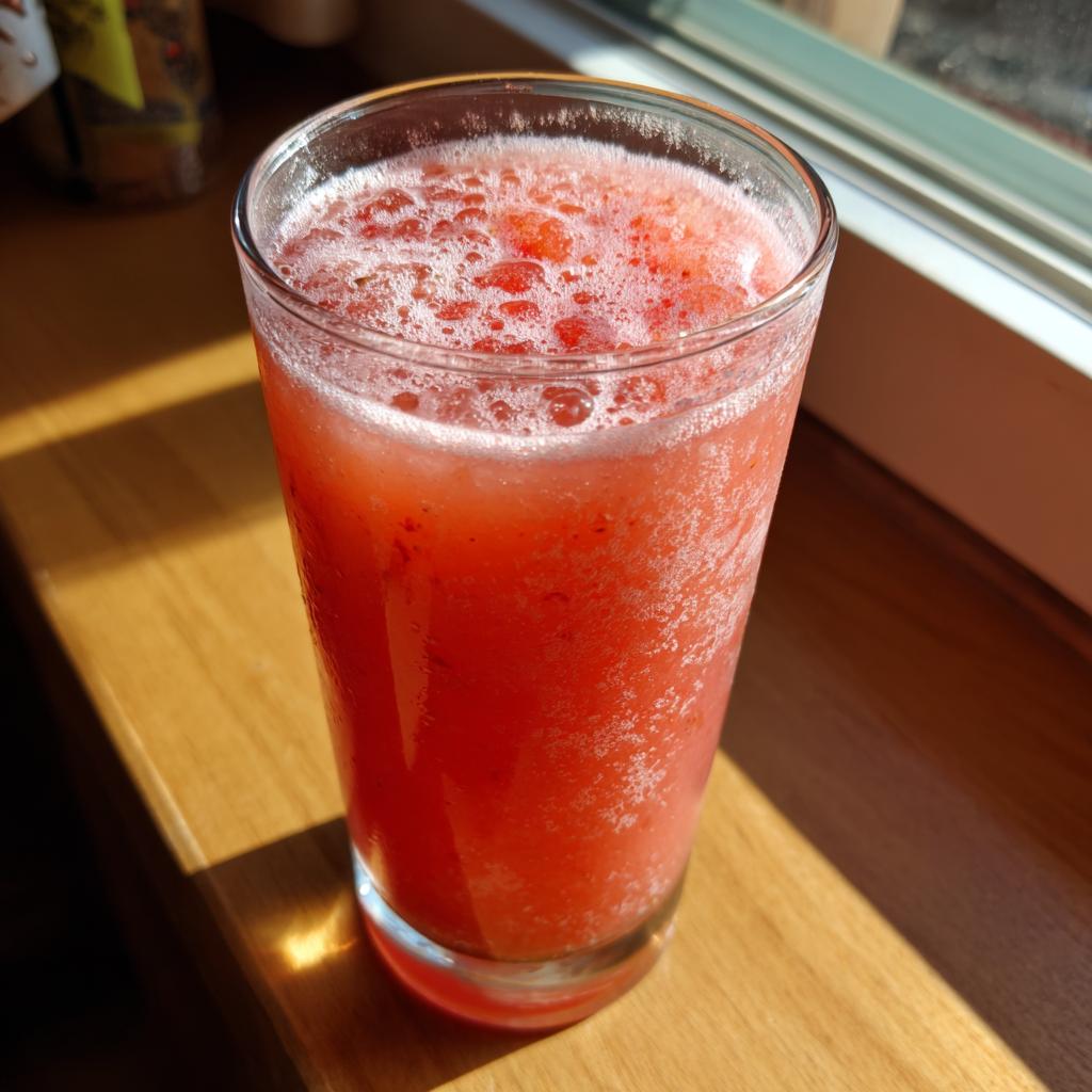 A tall glass filled with vibrant pink strawberry acai peach juice, with a frothy top, sitting on a wooden surface.