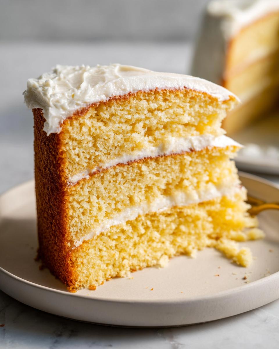 Close-up of a slice of vanilla cake with white frosting on a plate, showing moist layers.