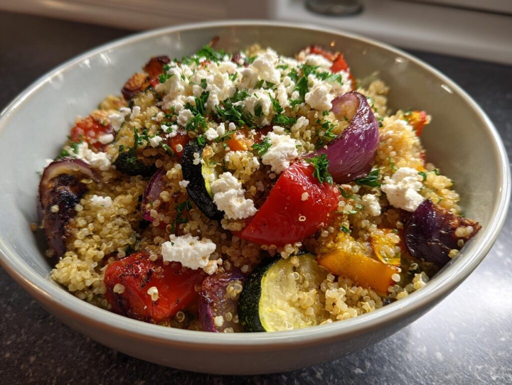 Bowl of quinoa salad with roasted vegetables and crumbled feta cheese garnished with herbs.