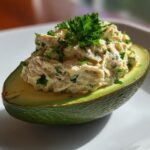 Fresh avocado half filled with creamy tuna salad garnished with parsley on white plate