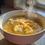 White bowl filled with creamy cauliflower soup with cheddar light, topped with cauliflower florets and shredded cheese.