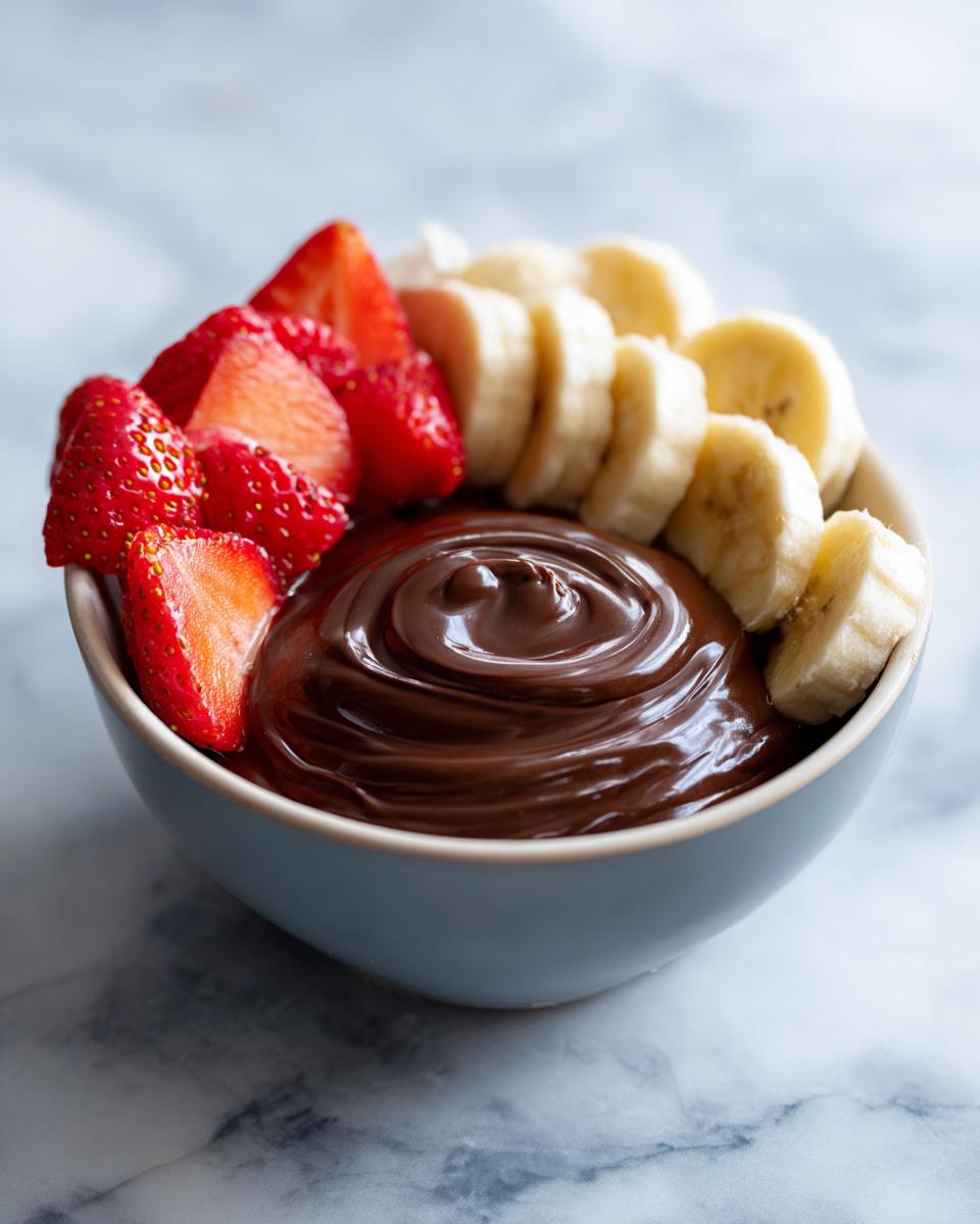 Bowl of creamy chocolate fruit dip with fresh sliced strawberries and bananas on the side.