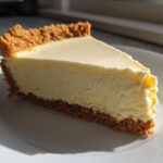 Close-up of a creamy slice of cheesecake with a golden graham cracker crust on a white plate