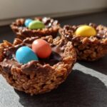 Close-up of Easter crispy chocolate nests filled with colorful candy eggs on a dark surface.