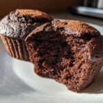 Two easy chocolate muffins on a white plate, one with a bite taken out showing moist texture.