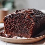 Close-up of a moist chocolate loaf cake with shiny chocolate glaze on a beige plate.