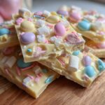 Close-up of white chocolate bark Easter pieces with pastel candies and marshmallows on wooden board.